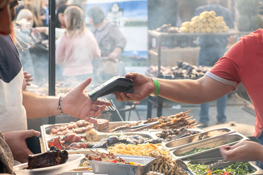Two people with identifying custom wristbands pass a payment scanning device across serving trays of food at an event